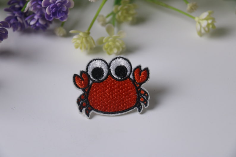 Op de afbeelding: Rode geborduurde strijk-op patch van een cartoon krab met grote witte ogen en zwarte pupillen.