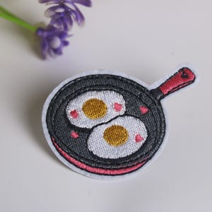 May include: Embroidered patch featuring a cartoon frying pan with two sunny-side-up eggs. The pan is black with a red handle and pink accents. The eggs have yellow yolks and white whites, with small pink hearts.