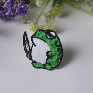 Könnte beinhalten: Gestickter Aufnäher mit einer grünen Frosch, der ein Messer hält. Der Frosch ist weiß mit schwarzen Umrissen und grünen Flecken. Das Messer ist schwarz mit einem weißen Griff.