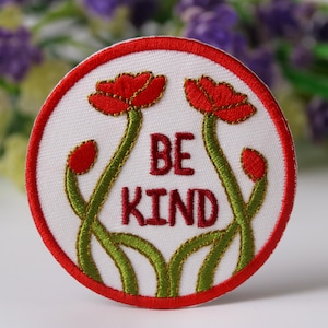Może przedstawiać: Biała naszywka haftowana z czerwoną obwódką. Naszywka przedstawia dwa czerwone kwiaty z zielonymi łodygami i liśćmi. Tekst "BE KIND" jest wyszyty na naszywce na czerwono.