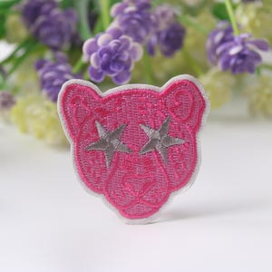 Può includere: Toppa ricamata rosa da stirare con contorno bianco e due occhi a forma di stella argentata.