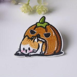 May include: An embroidered iron-on patch featuring a cartoon cat hiding under a smiling pumpkin. The pumpkin is orange with a green stem and the cat is white with orange and brown markings.