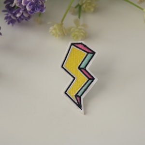 May include: A yellow lightning bolt shaped iron-on patch with pink and blue accents. The patch has a 3D effect.