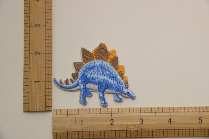 Pu&ograve; includere: Toppa ricamata blu e marrone da stirare a forma di dinosauro con una schiena spinosa.
