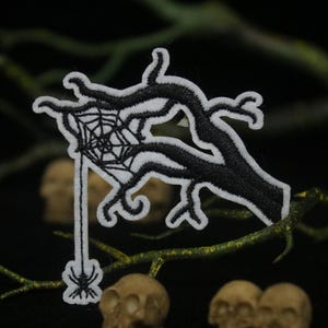 May include: Black and white embroidered patch of a tree branch with a spider web and a spider hanging from it.