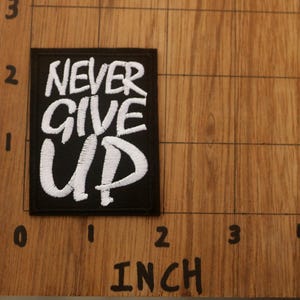 K&ouml;nnte beinhalten: Schwarzer rechteckiger Aufn&auml;her mit den Worten "NEVER GIVE UP" in Wei&szlig; gestickt. Der Aufn&auml;her ist etwa 5 cm breit und 5 cm hoch. Der Aufn&auml;her liegt auf einer Holzoberfl&auml;che.