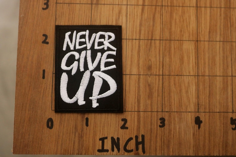 K&ouml;nnte beinhalten: Schwarzer rechteckiger Aufn&auml;her mit den Worten "NEVER GIVE UP" in Wei&szlig; gestickt. Der Aufn&auml;her ist etwa 5 cm breit und 5 cm hoch. Der Aufn&auml;her liegt auf einer Holzoberfl&auml;che.