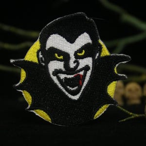 Puede incluir: Un parche bordado en blanco y negro de un vampiro con fondo amarillo. El vampiro tiene una gran sonrisa con una gota roja de sangre en el labio.