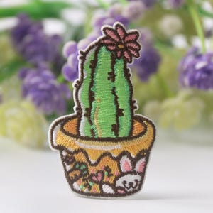 Puede incluir: Un cactus verde con una flor rosa en la parte superior, plantado en una maceta amarilla con un borde marrón y un diseño de conejo blanco.