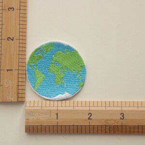 Pode incluir: Um remendo bordado que representa a Terra em azul e verde, com costuras brancas ao redor das bordas. O remendo mede aproximadamente 5 cm de di&acirc;metro.