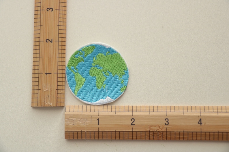 Pode incluir: Um remendo bordado que representa a Terra em azul e verde, com costuras brancas ao redor das bordas. O remendo mede aproximadamente 5 cm de di&acirc;metro.