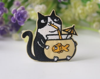 Op de afbeelding: Een zwart-witte kat met een wit gezicht en zwarte oren zit in een gele kom met een goudvis erin. De kat houdt een rietje in zijn mond en er is een gele paraplu achter hem.