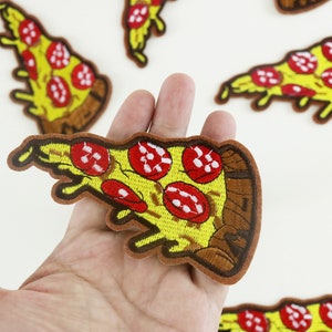 May include: A hand holding a yellow and brown embroidered iron-on patch of a pizza slice with red pepperoni and melted cheese.