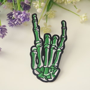 May include: Green and white embroidered skeleton hand making the rock and roll hand gesture. The patch is black and white with green stitching.