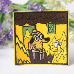Puede incluir: Parche bordado con un perro amarillo con un sombrero marrón sentado en una silla en una habitación envuelta en llamas. El perro sostiene una taza de café y mira al espectador con una expresión de sorpresa. El texto "This is fine" está escrito en la esquina inferior derecha del parche.