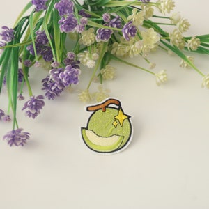 Peut inclure: &Eacute;cusson brod&eacute; repr&eacute;sentant un melon vert avec une tranche coup&eacute;e, une tige marron et une &eacute;toile jaune. L'&eacute;cusson a une bordure blanche. Des fleurs violettes et blanches avec des tiges vertes sont en arri&egrave;re-plan.