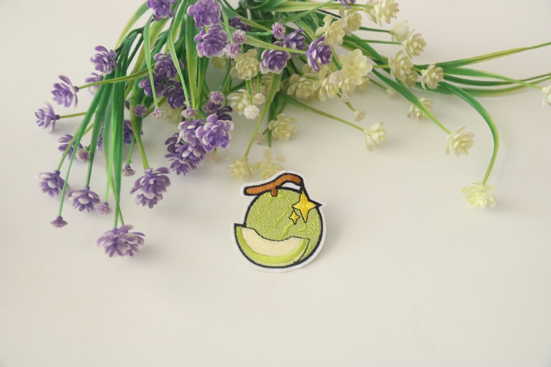 Peut inclure: &Eacute;cusson brod&eacute; repr&eacute;sentant un melon vert avec une tranche coup&eacute;e, une tige marron et une &eacute;toile jaune. L'&eacute;cusson a une bordure blanche. Des fleurs violettes et blanches avec des tiges vertes sont en arri&egrave;re-plan.