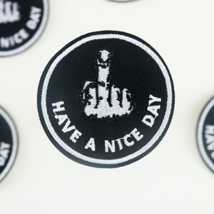 Può includere: Toppa ricamata in bianco e nero con un dito medio e il testo "Have a nice day".
