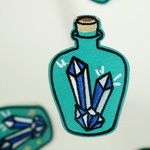 Puede incluir: Un parche bordado que representa una botella de vidrio azul turquesa con un tapón de corcho, que contiene dos formas de cristal azul y blanco.