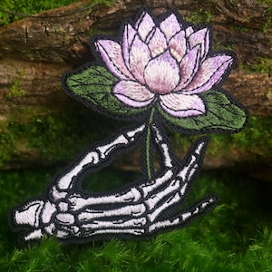 May include: An embroidered patch featuring a white and light purple lotus flower with green leaves held by a skeletal hand. The design is outlined in black thread, showcasing detailed stitching on the petals and the bones of the hand.