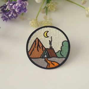 May include: Embroidered iron-on patch featuring a mountain, a small house with smoke coming from the chimney, a winding path, and a crescent moon in the sky. The patch is circular and has a black border.