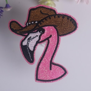 May include: An embroidered patch featuring a bright pink flamingo head wearing a brown cowboy hat. The flamingo has a black and white beak, and the entire design is outlined with a vibrant pink border that has a subtle glittery texture.