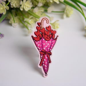 Pode incluir: Patch bordado com um design de guarda-chuva rosa preenchido com corações vermelhos. O guarda-chuva tem um padrão de bolinhas e um laço vermelho. O patch é para roupas ou acessórios.