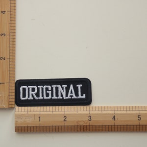 May include: Black fabric patch with white embroidered text that reads "ORIGINAL". The patch is measured against a wooden ruler.