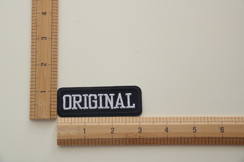 May include: Black fabric patch with white embroidered text that reads "ORIGINAL". The patch is measured against a wooden ruler.