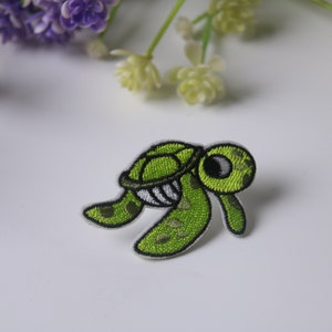 Può includere: Toppa ricamata di una tartaruga dei cartoni animati nei toni del verde, nero e bianco. La tartaruga ha un guscio dettagliato, una bocca sorridente e un occhio grande. La toppa è per abbigliamento o accessori.