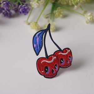 May include: Embroidered iron-on patch of two red cherries with white and purple skull faces and a blue stem with a purple and blue leaf.