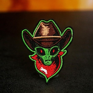 Könnte beinhalten: Gestickter Aufnäher mit einem grünen Alien, das einen braunen Cowboyhut und ein rotes Halstuch trägt. Das Alien hat rote Augen und ist grün umrandet. Der Aufnäher ist detailliert gestickt.