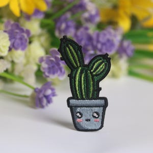May include: Embroidered cactus patch in a gray pot with a smiling face and pink cheeks. The cactus is green with dark green stripes and black outline. The patch is approximately 2 inches tall.