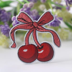 May include: A red embroidered cherry patch with a pink bow. The patch is perfect for adding a touch of whimsy to clothing, bags, or other accessories.
