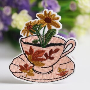 May include: Embroidered patch featuring a pink teacup and saucer with a floral design. The teacup holds yellow and brown flowers with green stems. Autumn leaves are embroidered on the cup and saucer. The patch has a white border.