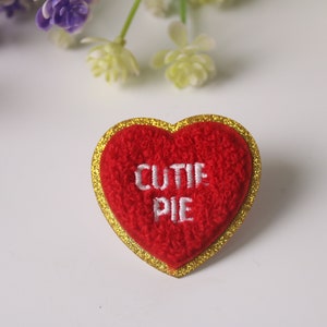 Puede incluir: Parche de fieltro rojo en forma de corazón con un borde de purpurina dorada. El parche tiene bordado blanco que dice "CUTIE PIE".