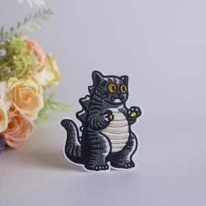 Peut inclure: Un patch brod&eacute; noir et blanc repr&eacute;sentant un chat aux grands yeux, ressemblant &agrave; un monstre, avec une bande jaune sur le dos.