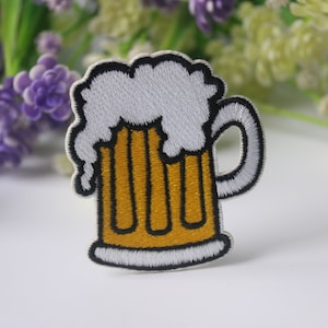 Op de afbeelding: Geborduurde strijkpatch van een bierglas met wit schuim en geel bier erin. Het glas heeft een zwarte omtrek.