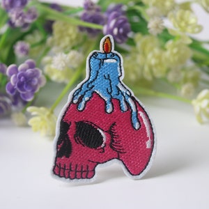 Puede incluir: Un parche bordado para planchar que presenta una calavera rosa con una vela azul derritiéndose en la parte superior. La vela tiene una llama amarilla.