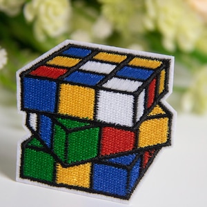 May include: Embroidered patch featuring a colorful Rubik's Cube design. The patch showcases the classic puzzle with its iconic arrangement of red, blue, yellow, green, white, and orange squares, outlined in black. This patch is ideal for adding to clothing or accessories.