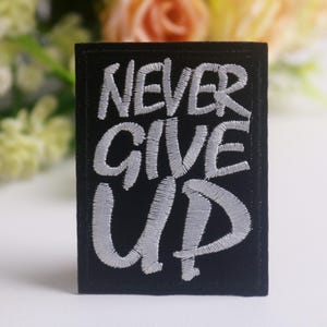 May include: Black rectangular patch with the embroidered words "NEVER GIVE UP" in white. The patch is set against a blurred background of flowers in shades of yellow, pink, and green.