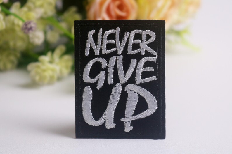 K&ouml;nnte beinhalten: Schwarzer rechteckiger Aufn&auml;her mit den gestickten Worten "NEVER GIVE UP" in Wei&szlig;. Der Aufn&auml;her ist vor einem verschwommenen Hintergrund aus Blumen in Gelb-, Rosa- und Gr&uuml;nt&ouml;nen platziert.