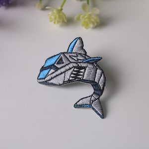 May include: A gray and blue embroidered iron-on patch of a stylized shark. The shark is facing left and has a blue fin and tail.