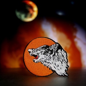 Op de afbeelding: Een witte geborduurde wolfskop patch met een open mond en scherpe tanden, op een fel oranje ronde achtergrond.