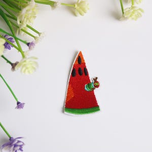 May include: Embroidered watermelon slice patch with a green and red color scheme, featuring a small caterpillar. The patch has black seeds and a white border. Decorative floral elements are in the background.