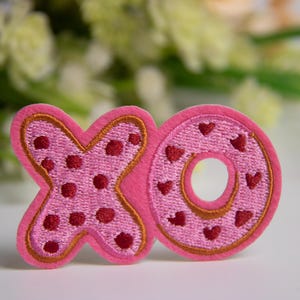 May include: A pink embroidered "XO" patch. The "X" is decorated with red circles, and the "O" with red hearts. It has a gold border. The background is a blurred floral arrangement. The patch is suitable for clothing.