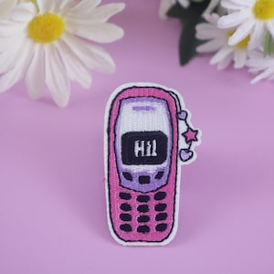 Puede incluir: Parche bordado con forma de teléfono móvil rosa con la palabra "Hi!" en la pantalla. El parche tiene un borde blanco y un adorno con estrellas y corazones. Las margaritas están en el fondo.