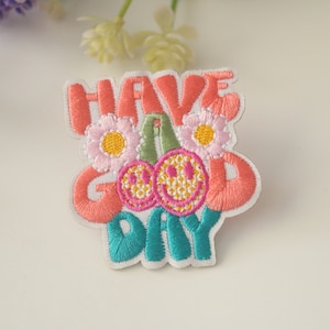 Puede incluir: Parche bordado para planchar con el texto "Have a Good Day" en letras rosa, verde y azul. El parche presenta dos caras sonrientes y dos flores rosas con centros amarillos.