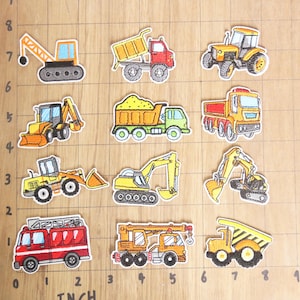 May include: Ten colorful iron-on patches featuring different types of construction vehicles, including a crane, dump truck, bulldozer, and excavator. The patches are arranged in a grid on a wooden surface with a ruler for scale.