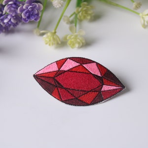 May include: A red and pink embroidered iron-on patch shaped like a gemstone. The patch is detailed with black stitching and has a geometric design.